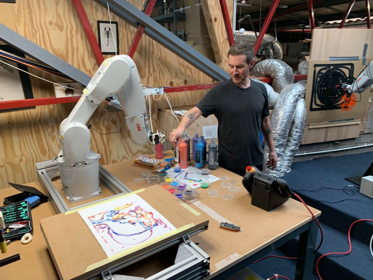 A robot arm plotting on a table in the studio while Andrew Melchior looks on.