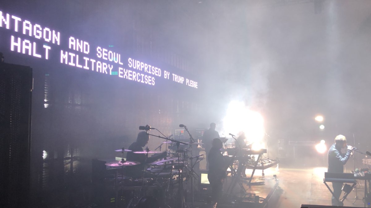 Massive Attack live — a stage LED wall running a scrolling news ticker in white on black.