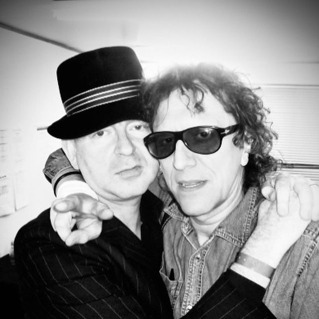 Black-and-white photograph of Mick Rock with a companion in a hat.