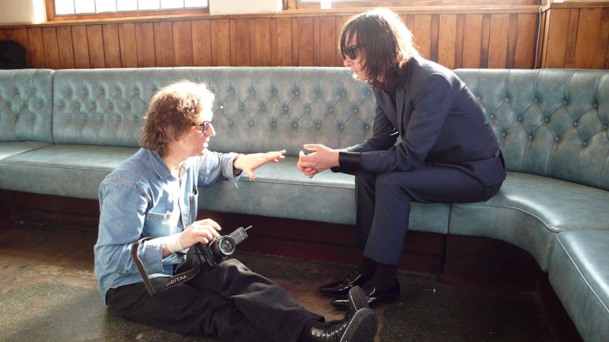 Mick Rock crouching to talk with a subject seated on a teal banquette.