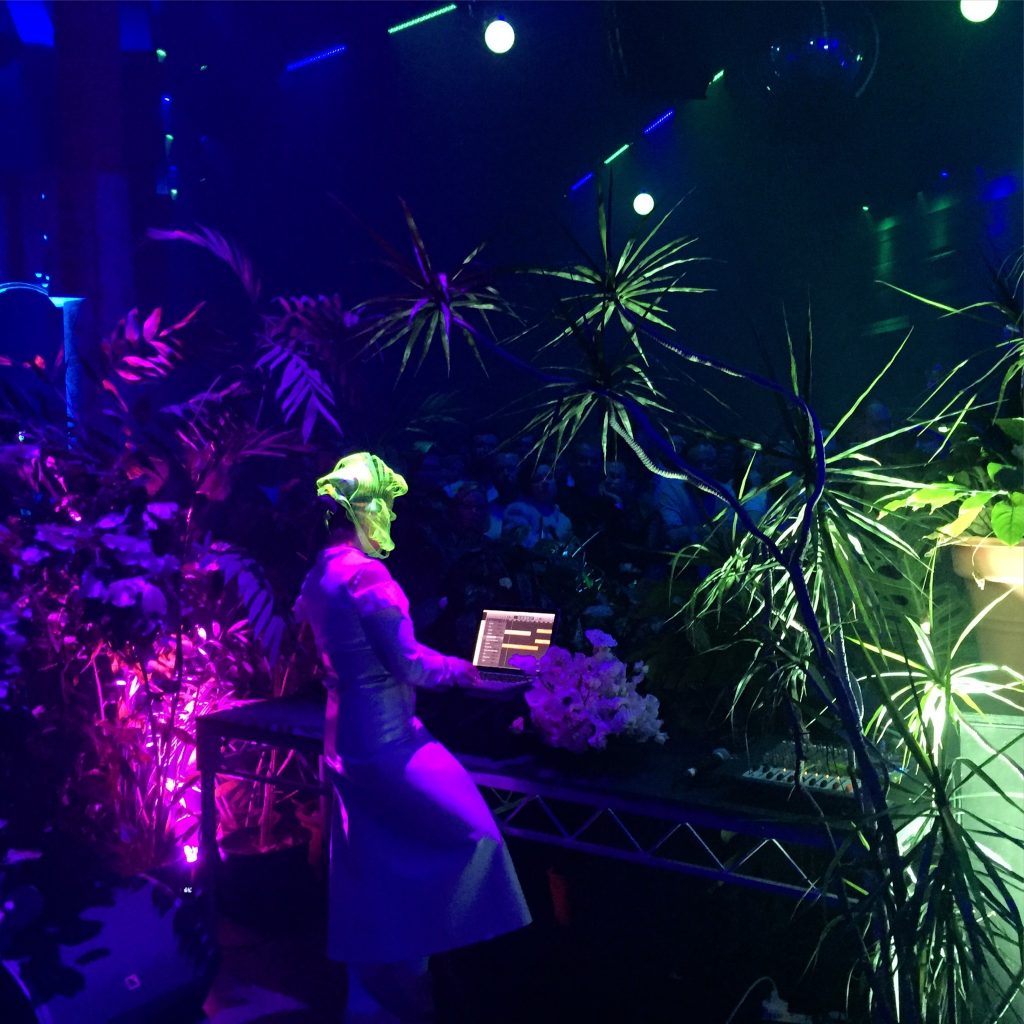 Masked performer at a laptop amid dense foliage under coloured stage light.