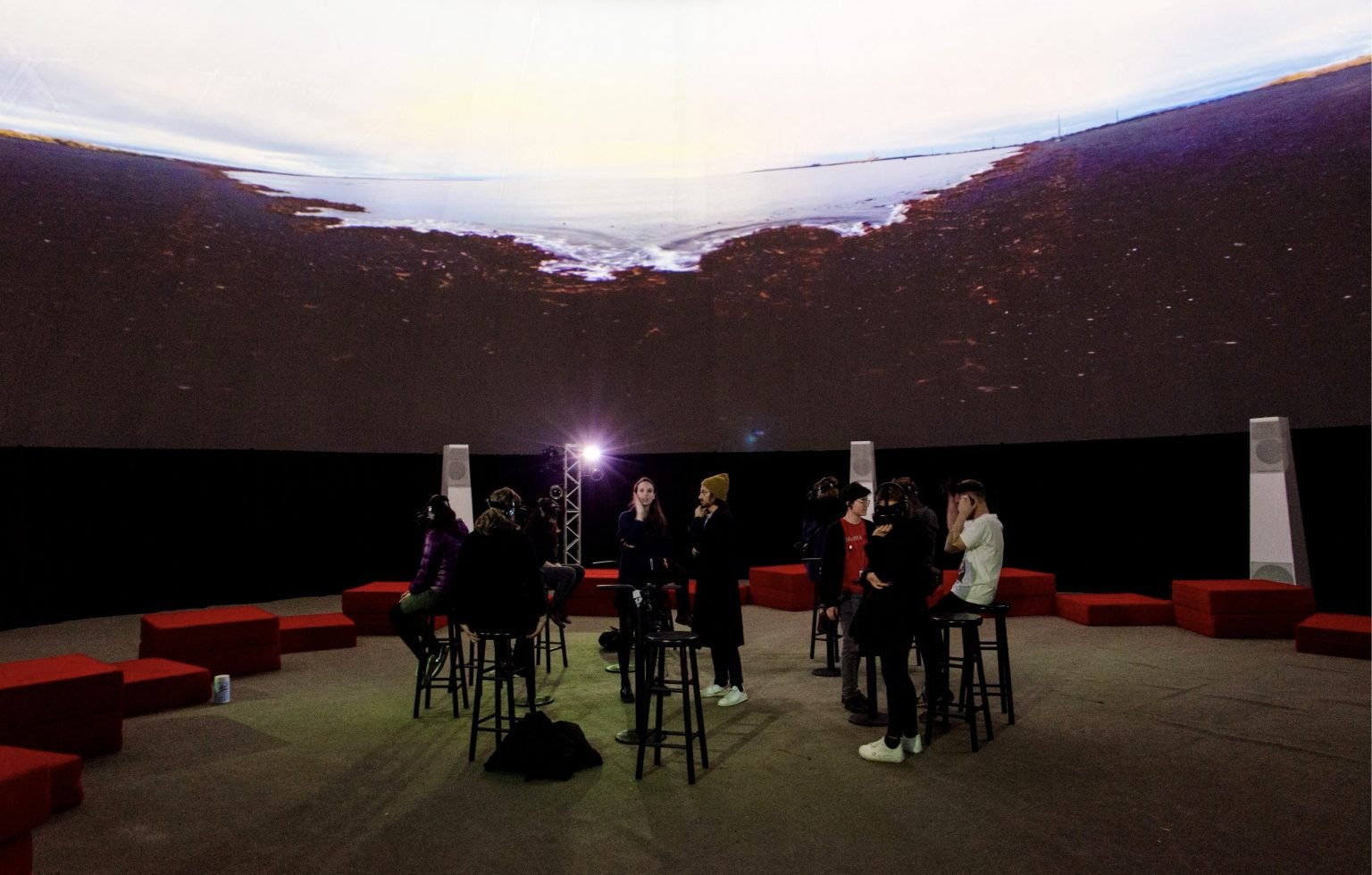 Audience inside a 360° immersive projection dome, seated among speakers as a vast landscape wraps overhead.